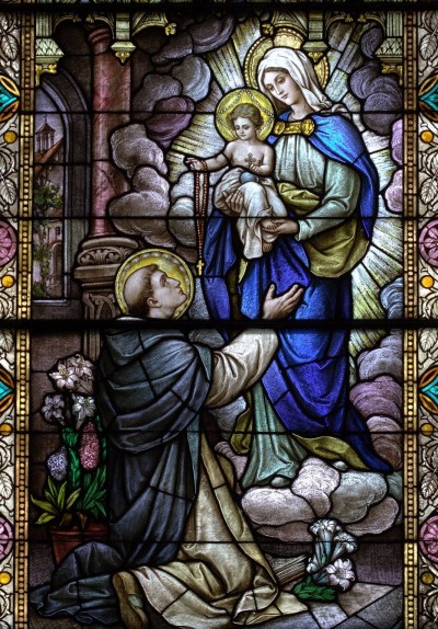 Na imagem do vitral, a Santíssima Virgem com o Menino Jesus entregam o Santo Rosário à São Domingos.